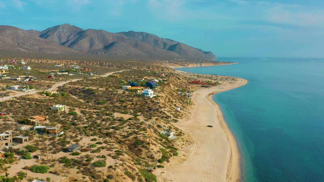 Frame at 0:02 shows El Sargento La Ventana, Mexico from above, with the shoreline sweeping toward the distant headland, homes set back in scrubland, blue water to the right, and no visible people.
