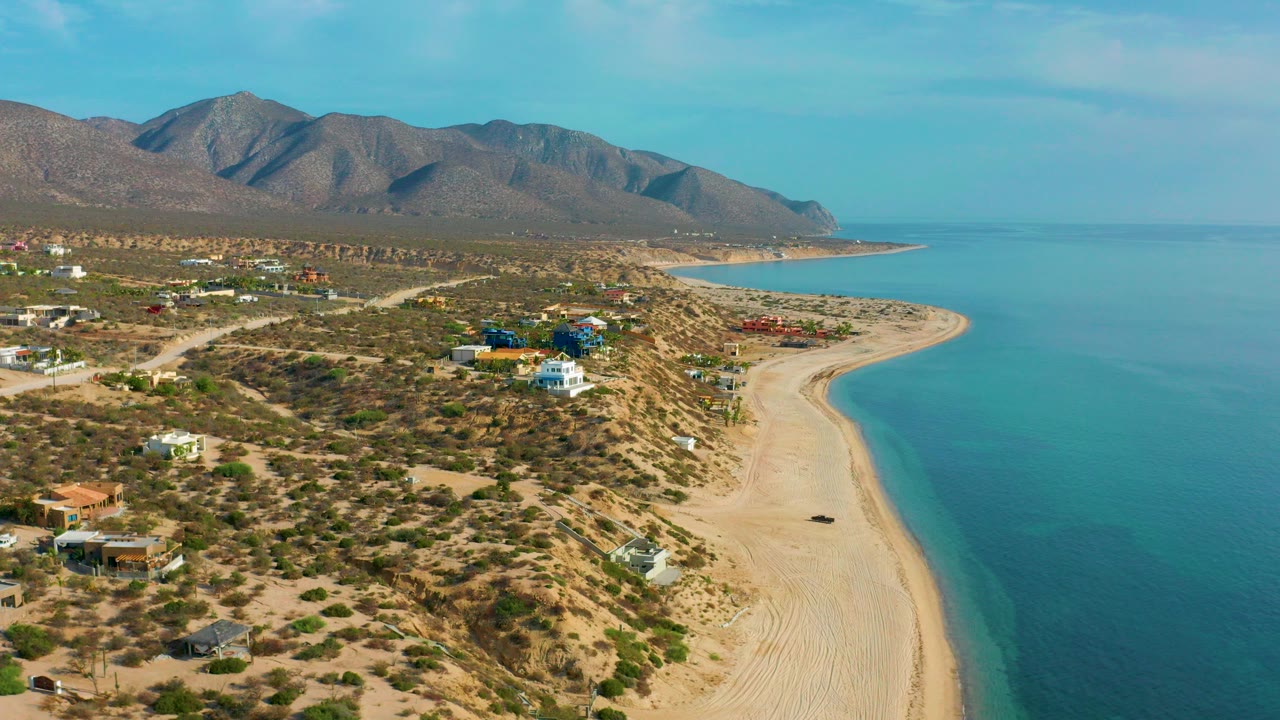 Aerial view over El Sargento La Ventana with scattered beachside homes, scrubby desert lots, a curving sandy shore, blue water, mountains in the distance, and no visible people.