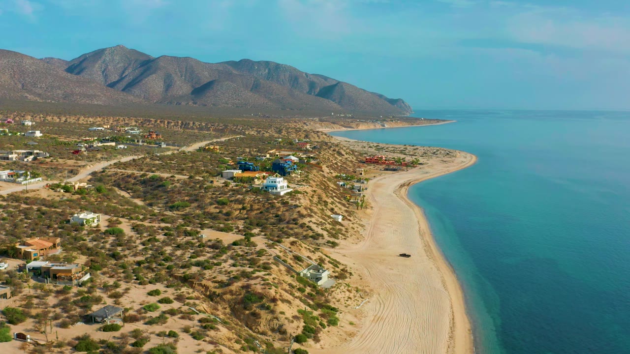 Wide aerial of a curved beach beside turquoise water and desert houses.