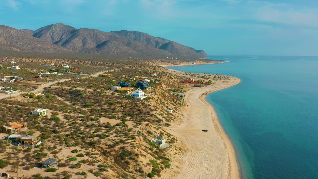 Aerial view over El Sargento La Ventana with clustered colored buildings near the shore, scrubland lots, a broad sandy beach, calm blue sea, mountains, and no visible people.