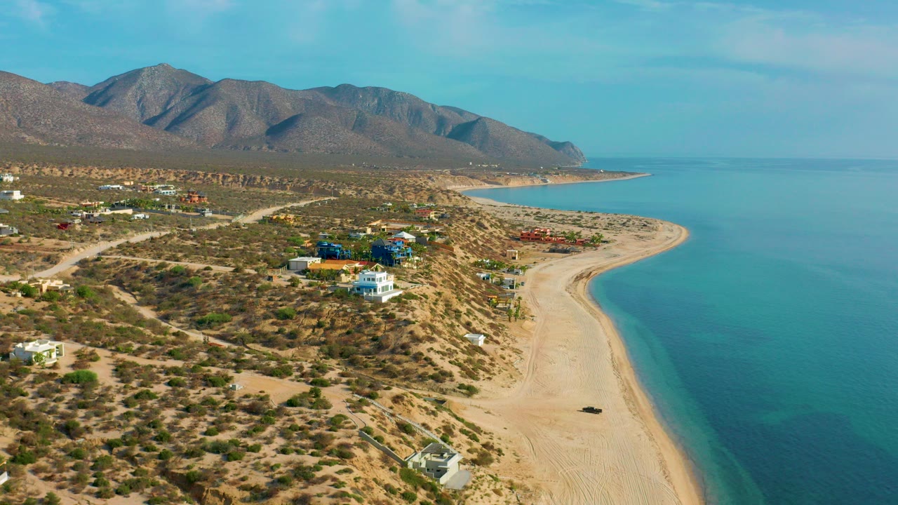 Aerial view over El Sargento La Ventana, Mexico, showing a curved sandy shoreline, turquoise water, scrubland roads, scattered buildings, brown mountains, and no visible people.