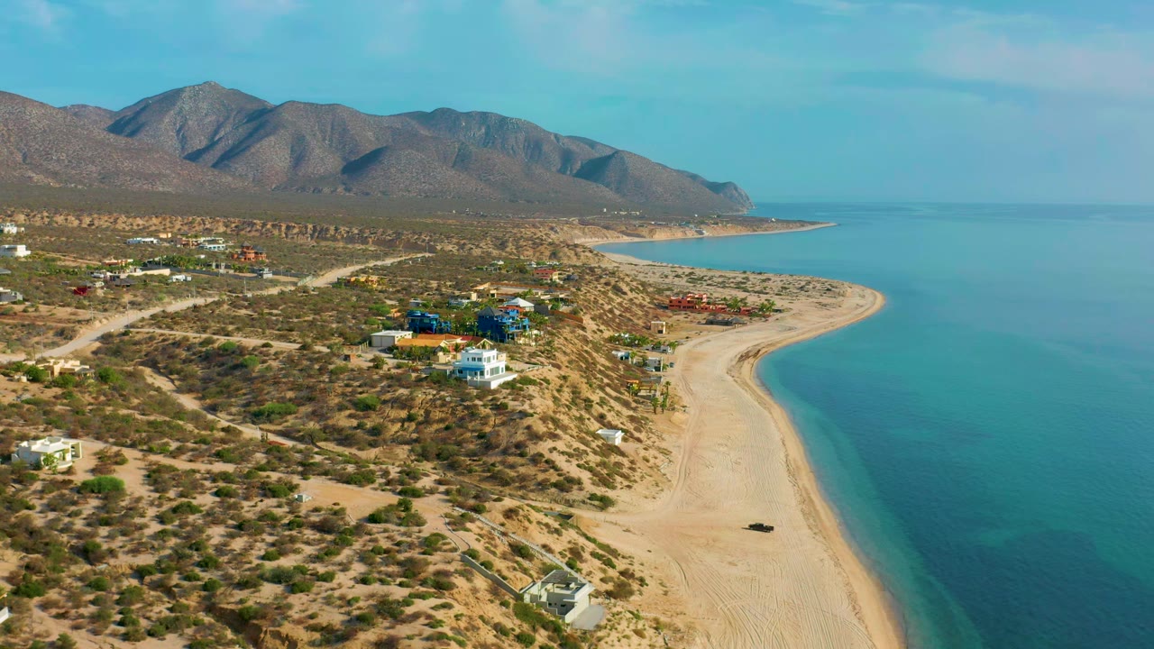 Wide aerial frame of El Sargento La Ventana, Mexico, with the beach running along the right edge, low buildings and dirt roads inland, mountains behind, and no visible people.