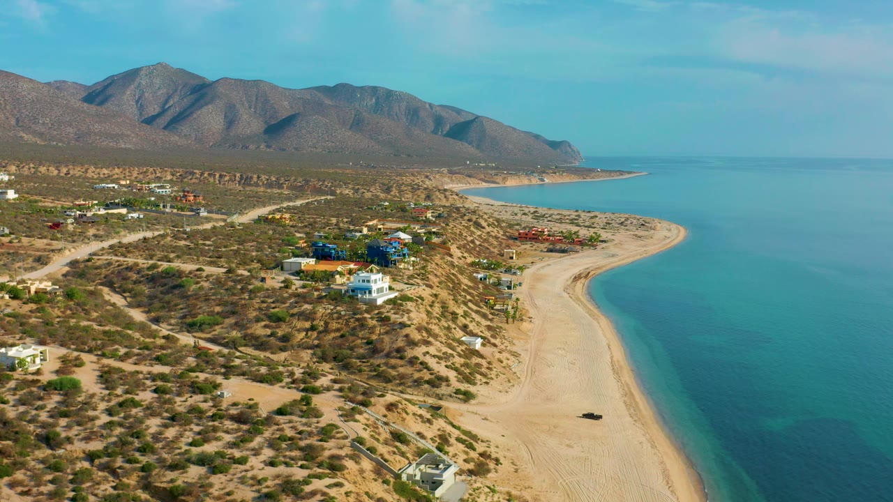 Elevated view over El Sargento La Ventana, Mexico, showing beach homes near the sand, a pale shoreline curving into calm blue water, mountain slopes, and no visible people.