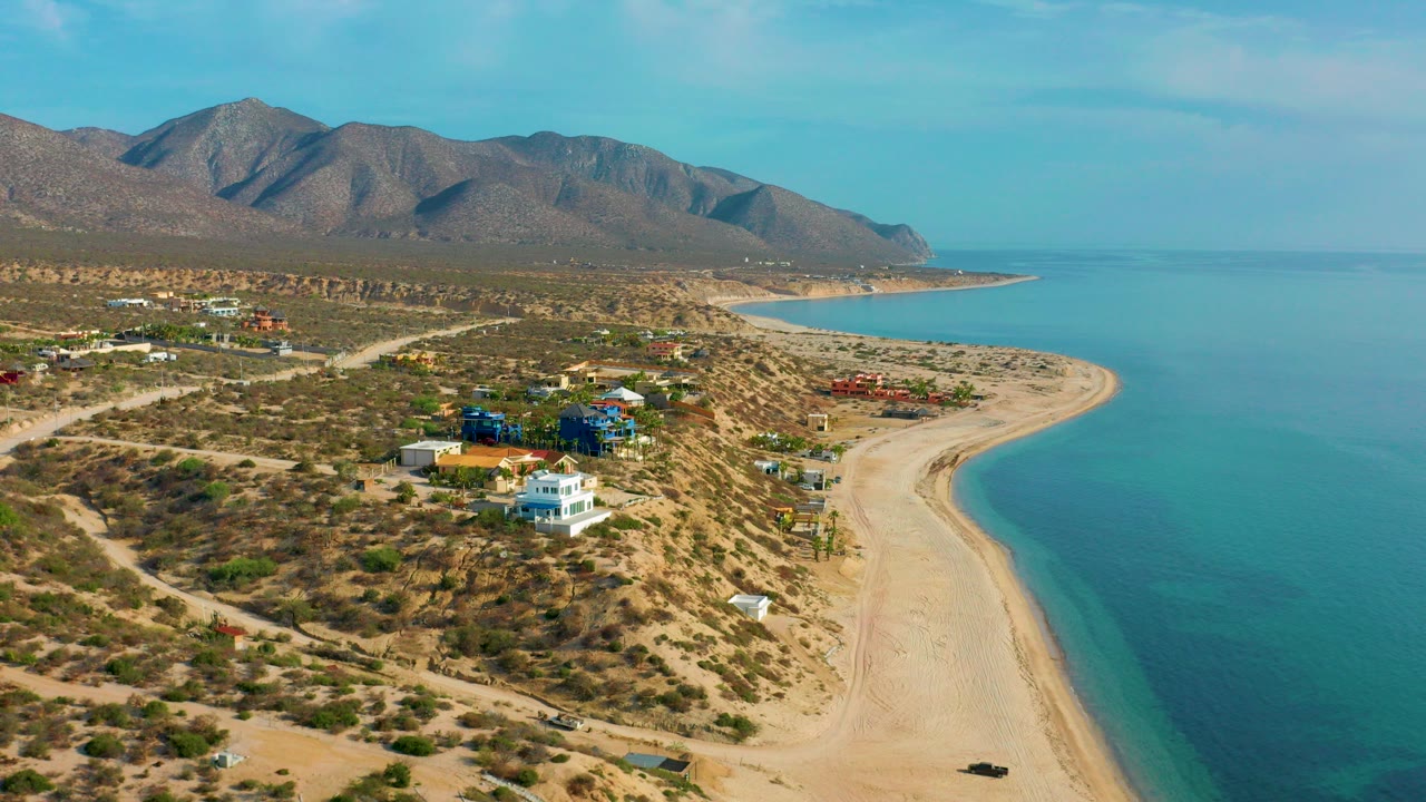 Opening aerial frame over El Sargento La Ventana, Mexico, showing scattered homes on dry coastal terrain, a pale beach curving beside blue water, mountains behind, and no visible people.