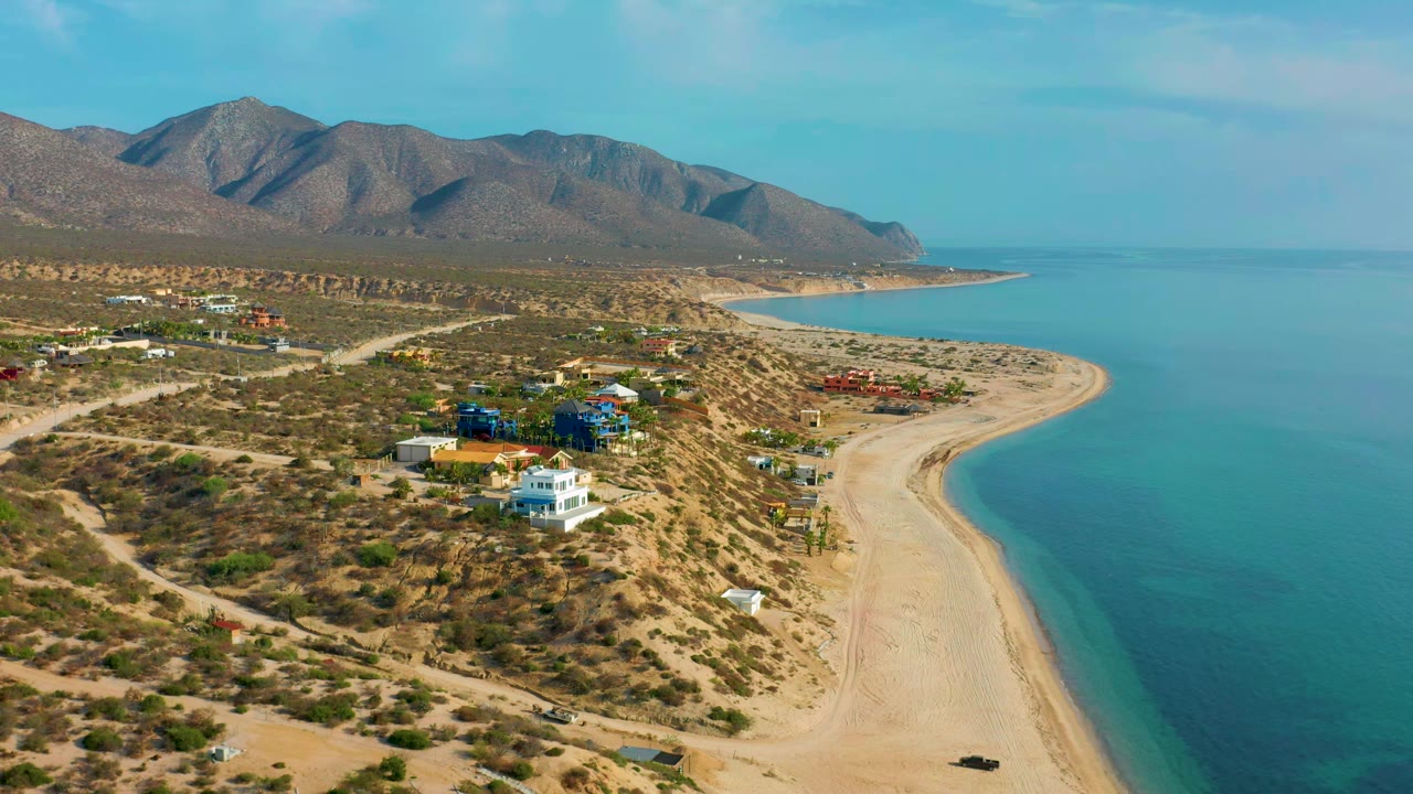 Aerial frame in El Sargento La Ventana, Mexico, with a white house and colorful homes in the lower frame, a curved sandy shoreline to the right, mountains inland, and no visible people.