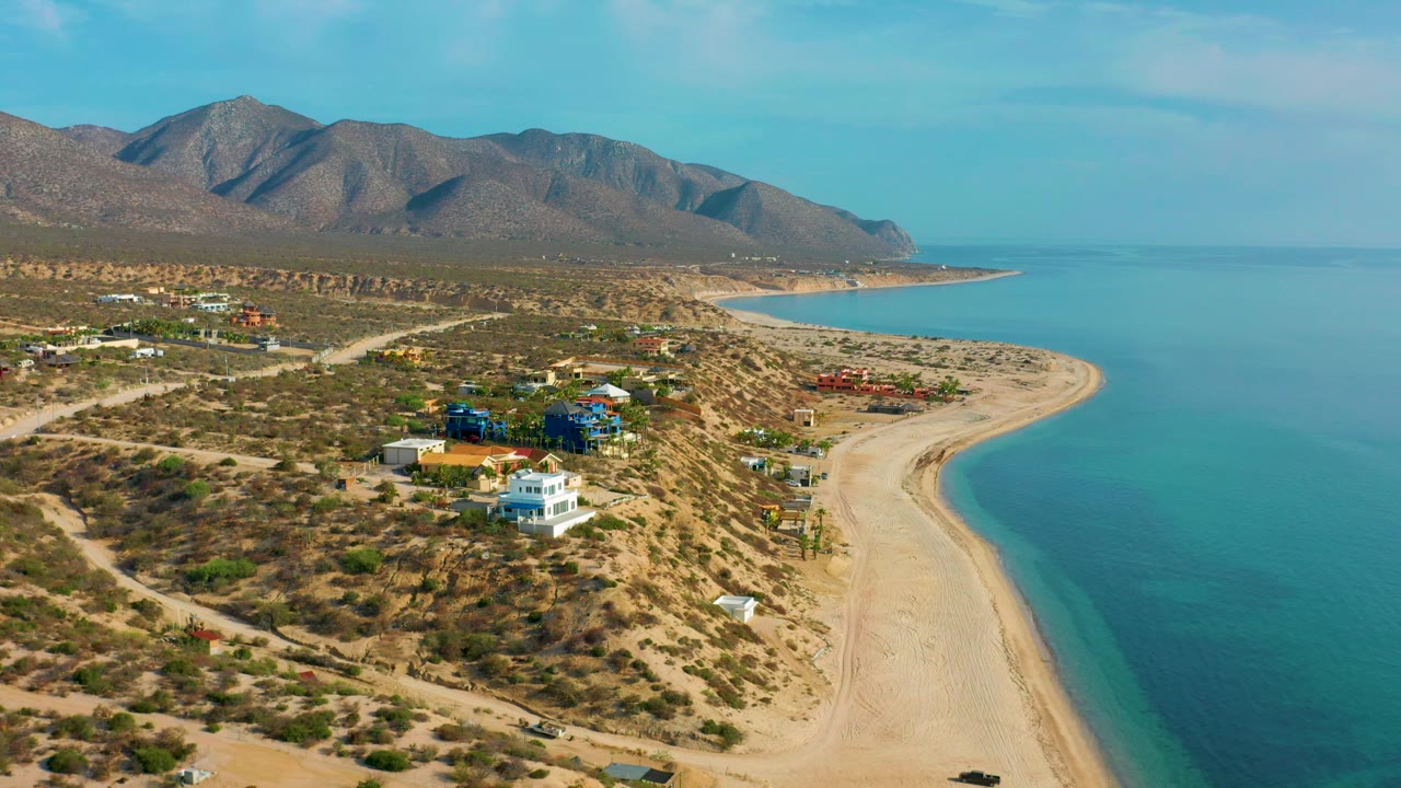 Aerial view over El Sargento La Ventana, Mexico, showing residential buildings across dry scrubland, a broad empty beach, clear blue-green water, distant mountains, and no visible people.
