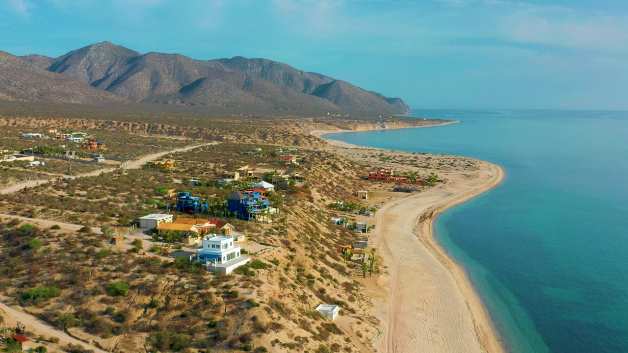 Aerial frame over El Sargento La Ventana, Mexico, with scattered coastal homes in the lower left, a pale beach curving along blue-green water, mountains beyond, and no visible people.