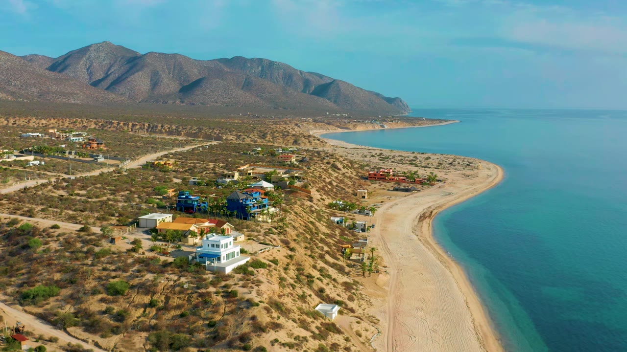 High coastal view with white and blue houses above a sandy shoreline and mountains.