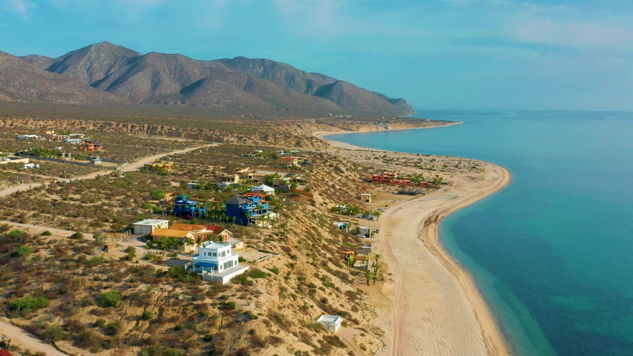 Aerial frame over the El Sargento La Ventana shoreline, with sparse houses on scrub-covered ground, a broad curved beach, blue water, mountain slopes across the background, and no visible people.