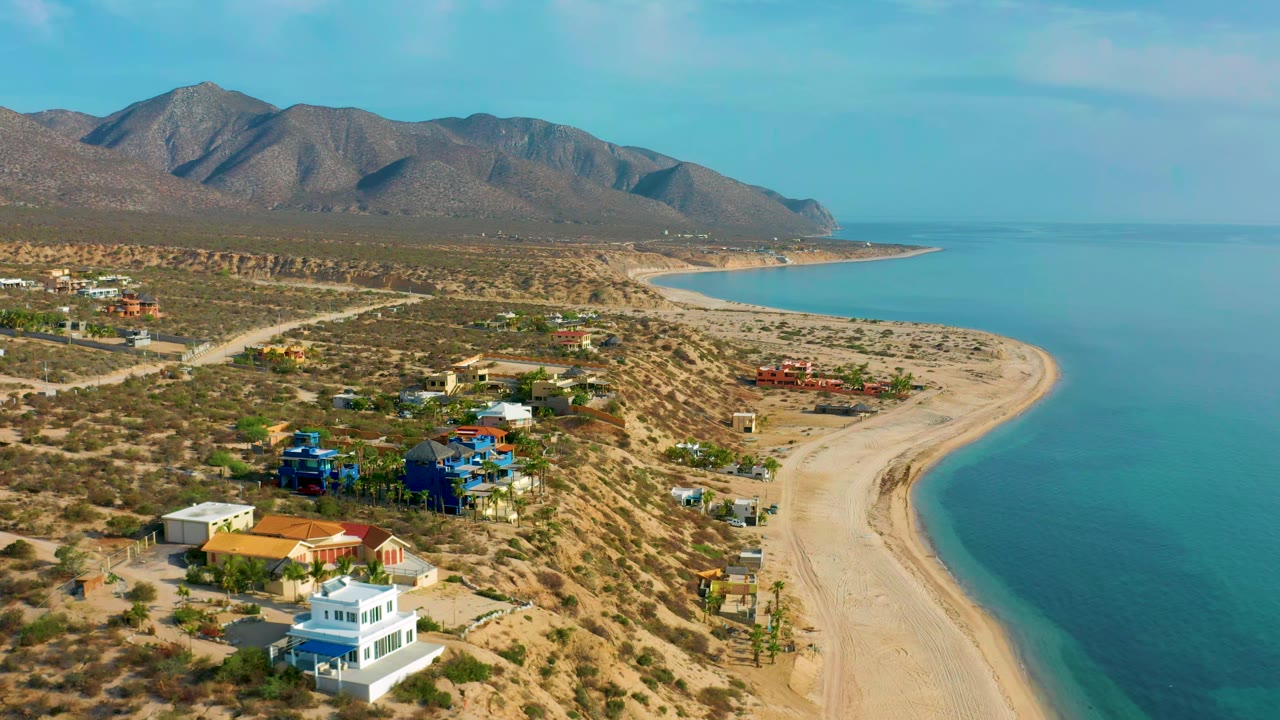 Coastal houses beside a curving beach with sea and mountains beyond.