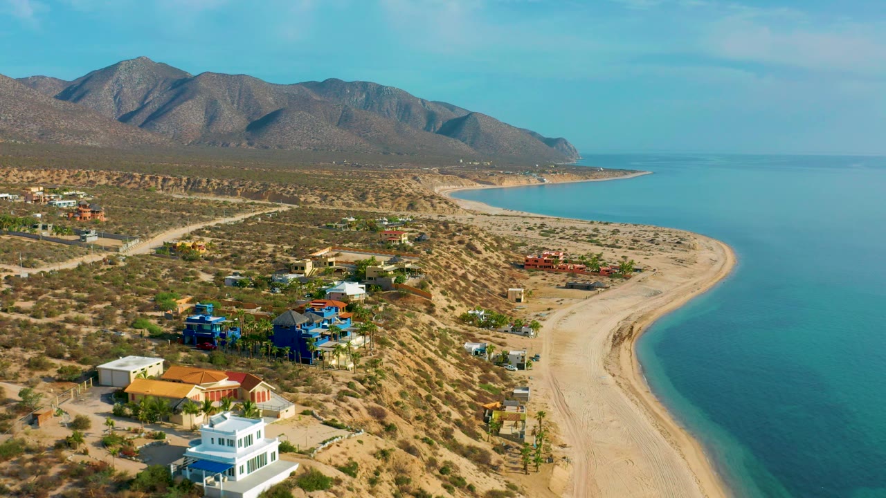 Aerial frame from El Sargento La Ventana, Mexico, showing colorful beachfront homes set in dry terrain, calm turquoise sea along a curved sandbar, distant mountains, and no visible people.