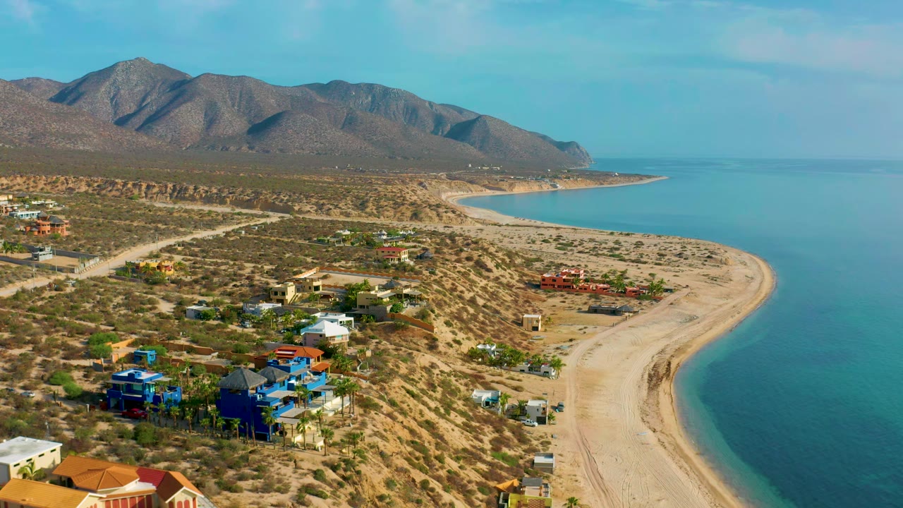 Aerial view in El Sargento La Ventana, Mexico, with colorful beachside homes in the lower frame, a pale sand curve, calm ocean, mountain ridge, and no visible people.