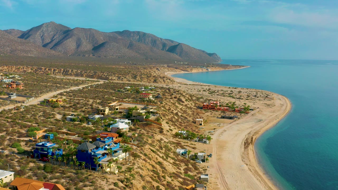 Drone perspective over El Sargento La Ventana, Mexico, showing blue and red-roofed houses near desert brush, a broad beach curve, turquoise water, mountains, and no visible people.