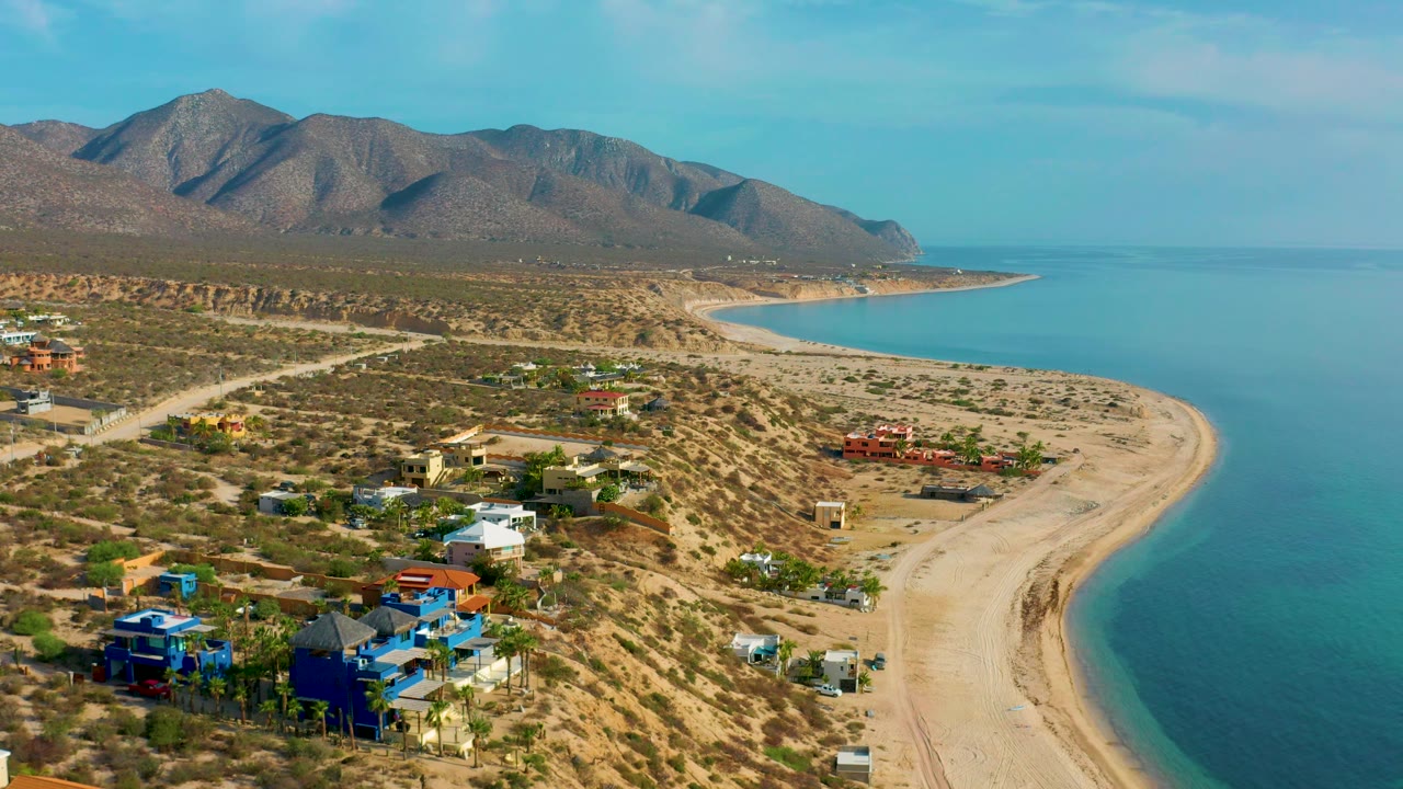 Aerial coastline with low houses, unpaved roads, sandy beach, ocean, and mountains.