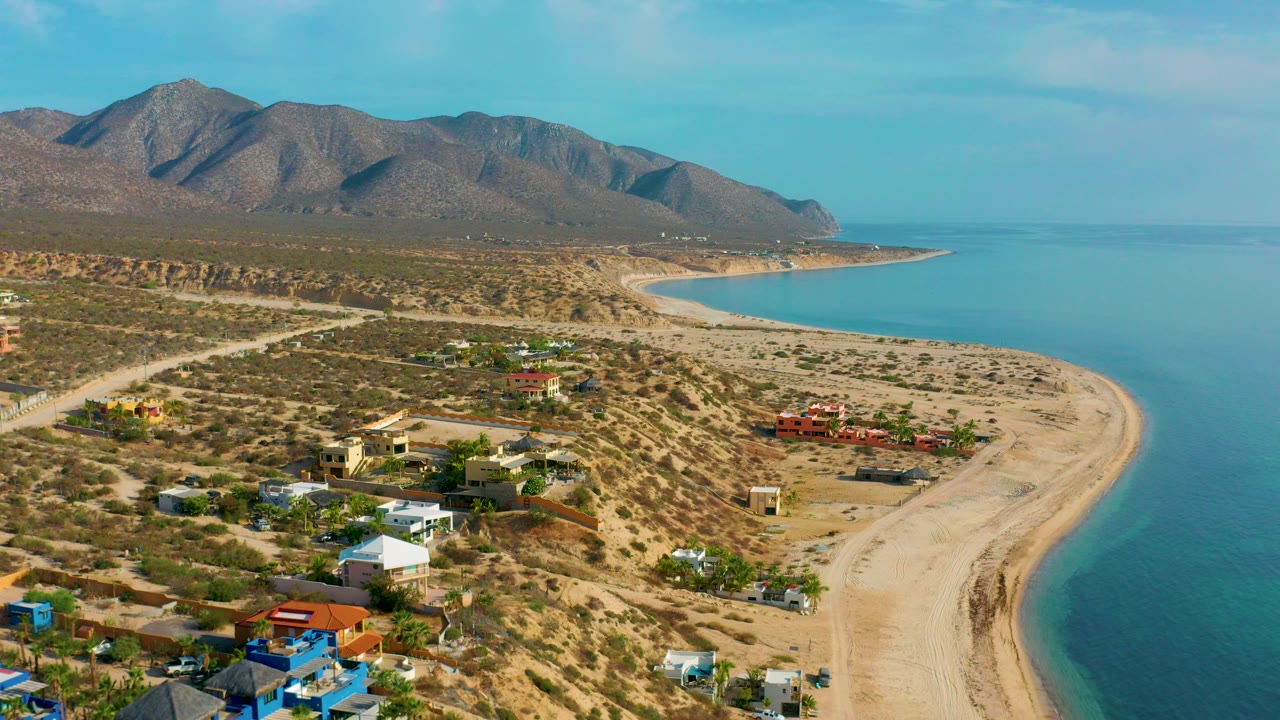 Early aerial frame over El Sargento La Ventana, Mexico, showing scattered homes near a sandy beach, calm blue water to the right, brown mountains in the distance, and no visible people.
