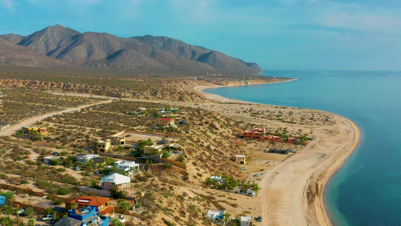 Aerial frame from El Sargento La Ventana, Mexico, showing a broader stretch of sandy shoreline, small homes set back from the beach, open blue water, mountain ridges, and no visible people.