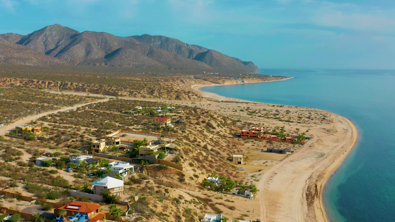 Aerial coastal village scene with beach, water, desert terrain, and mountains.