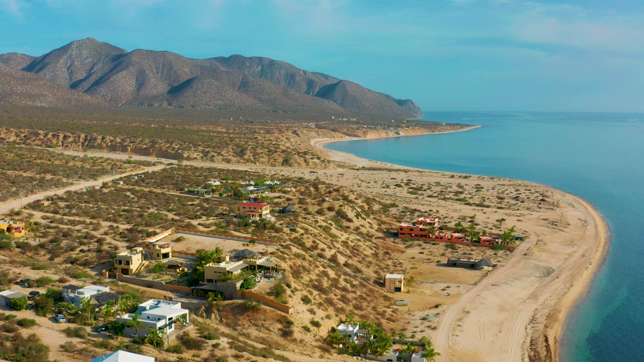 Aerial frame over El Sargento La Ventana, Mexico, showing private homes in sandy desert terrain, a curving beach, blue water, brown mountains, and no visible people.