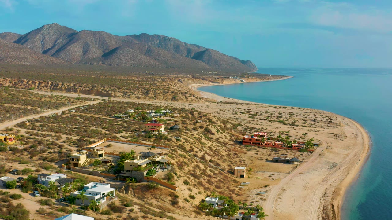 High view in El Sargento La Ventana, Mexico, with scattered private houses across arid lots, pale beach curving along blue sea, mountain ridge behind, and no visible people.