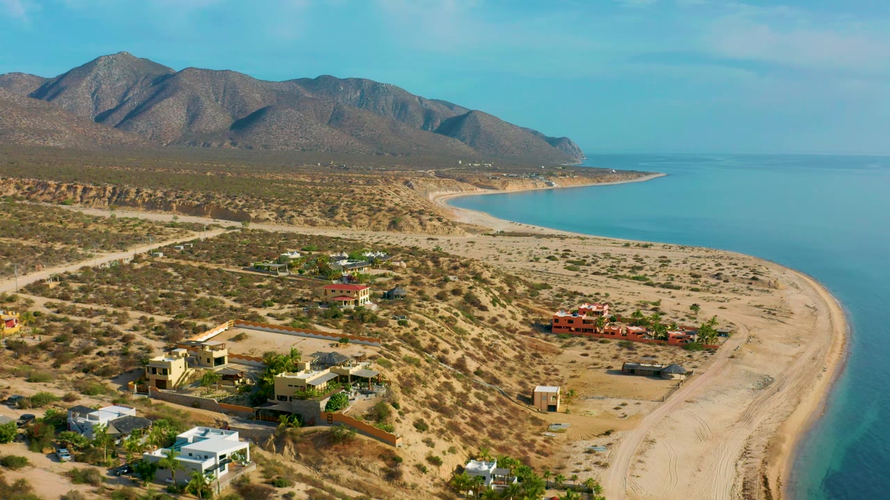 Wide aerial view of El Sargento La Ventana, Mexico, with desert lots and private homes in the foreground, a narrow sand beach beside blue water, distant mountains, and no visible people.