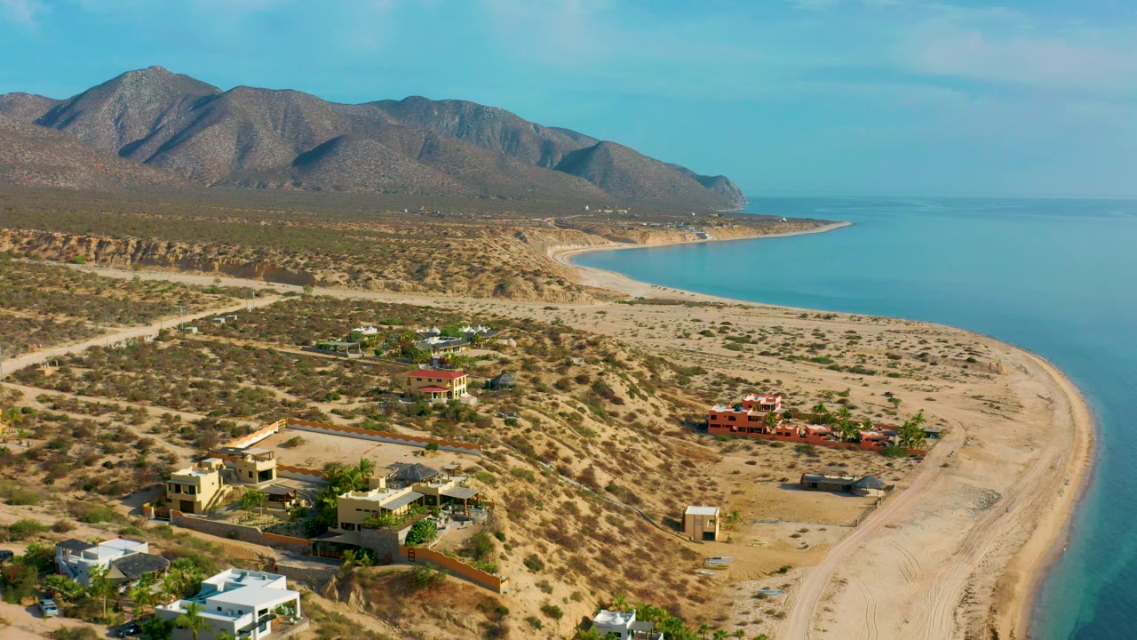 Aerial view of houses and scrubland above a beach with calm blue water.