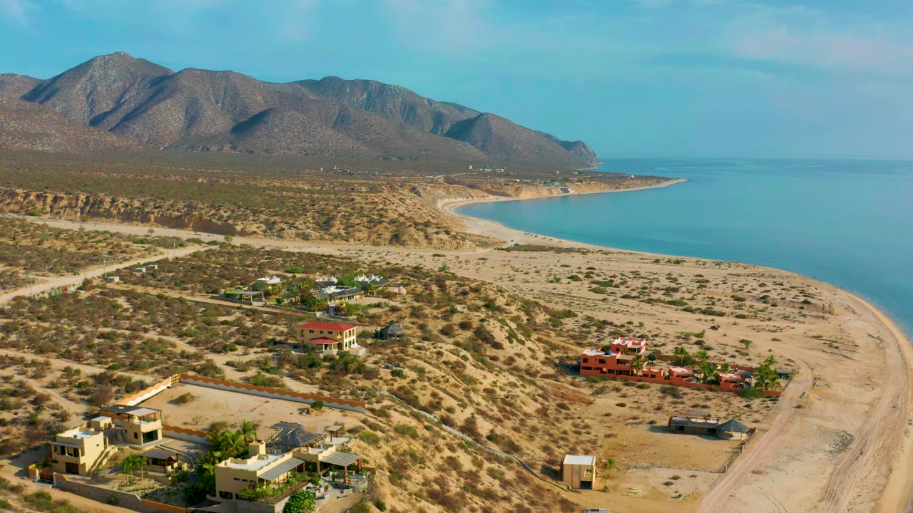 Wide aerial frame in El Sargento La Ventana, Mexico, showing modern houses near sandy lots, a pale beach curving along calm blue water, distant mountains, and no visible people.