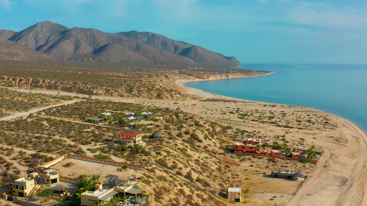 High view of El Sargento La Ventana, Mexico, with red and pale buildings in the foreground, desert scrub across the shore plain, blue sea at right, mountains beyond, and no visible people.