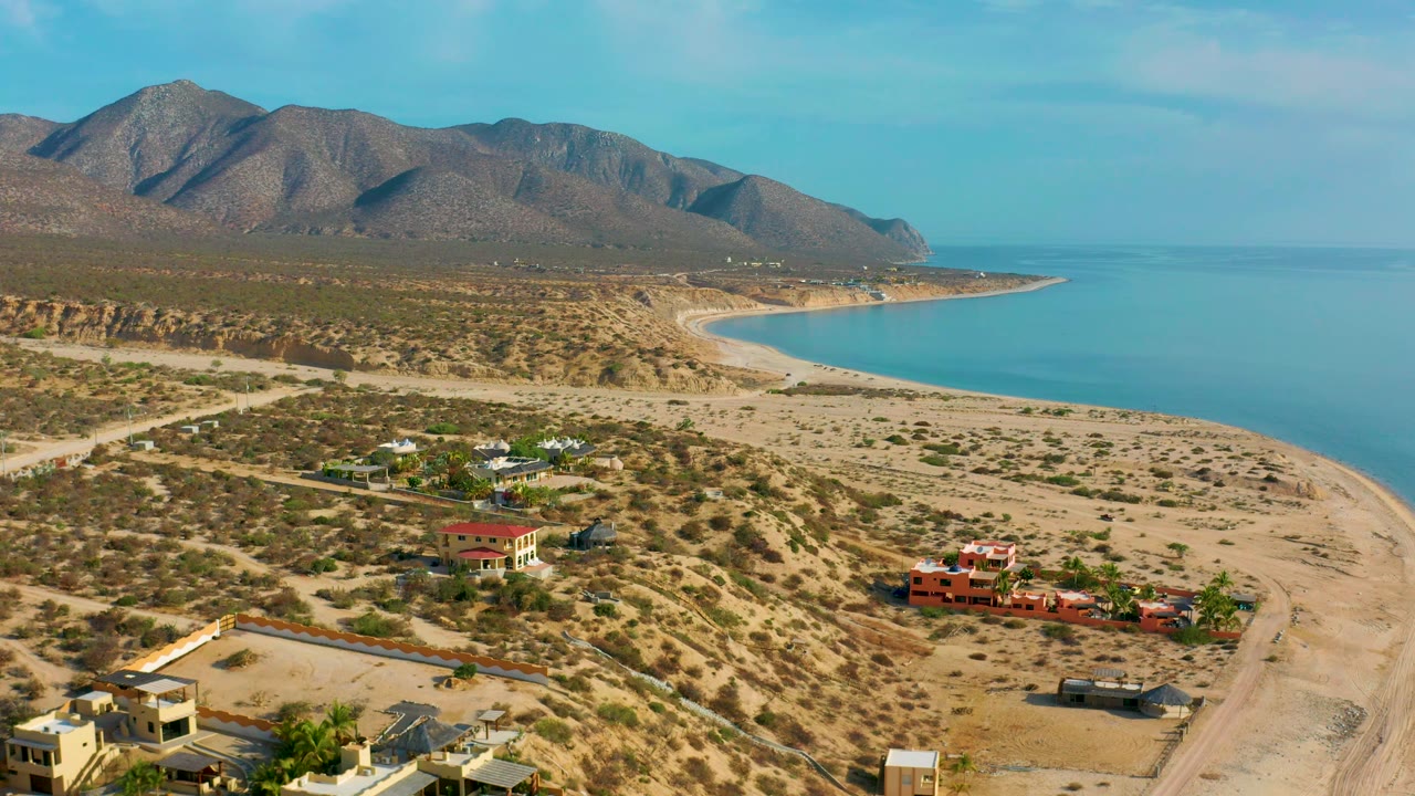Aerial frame of sparse houses, dirt tracks, beach, and mountain-backed water.