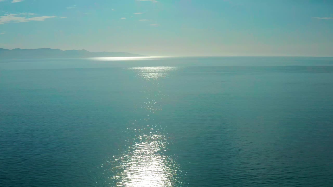 Opening frame of the El Sargento La Ventana, Mexico ocean view, with a bright vertical sun reflection on teal water, distant mountains at the horizon, pale sky, and no visible people.