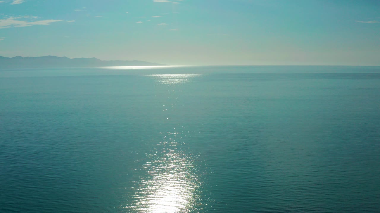 Blue sea with bright sunrise reflection and distant hills on the horizon.