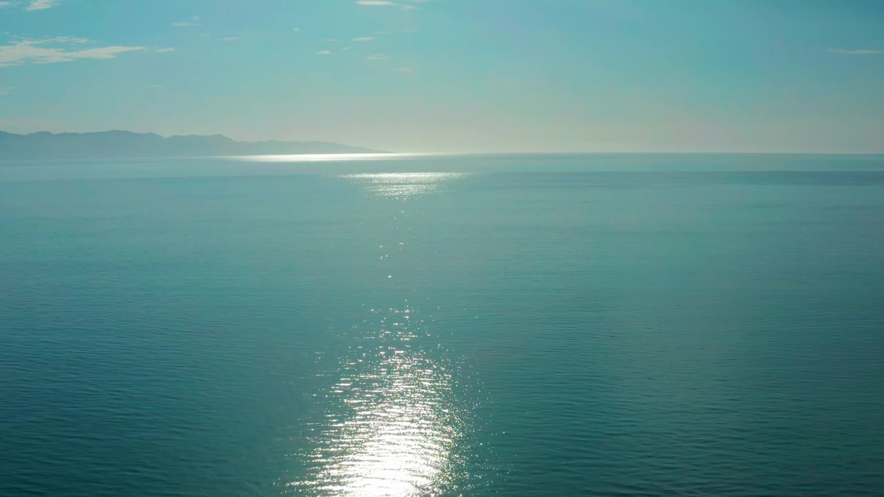 Early frame from El Sargento La Ventana, Mexico, showing open blue water, a compact bright reflection below the horizon, hazy landforms in the distance, light cloud streaks, and no visible people.