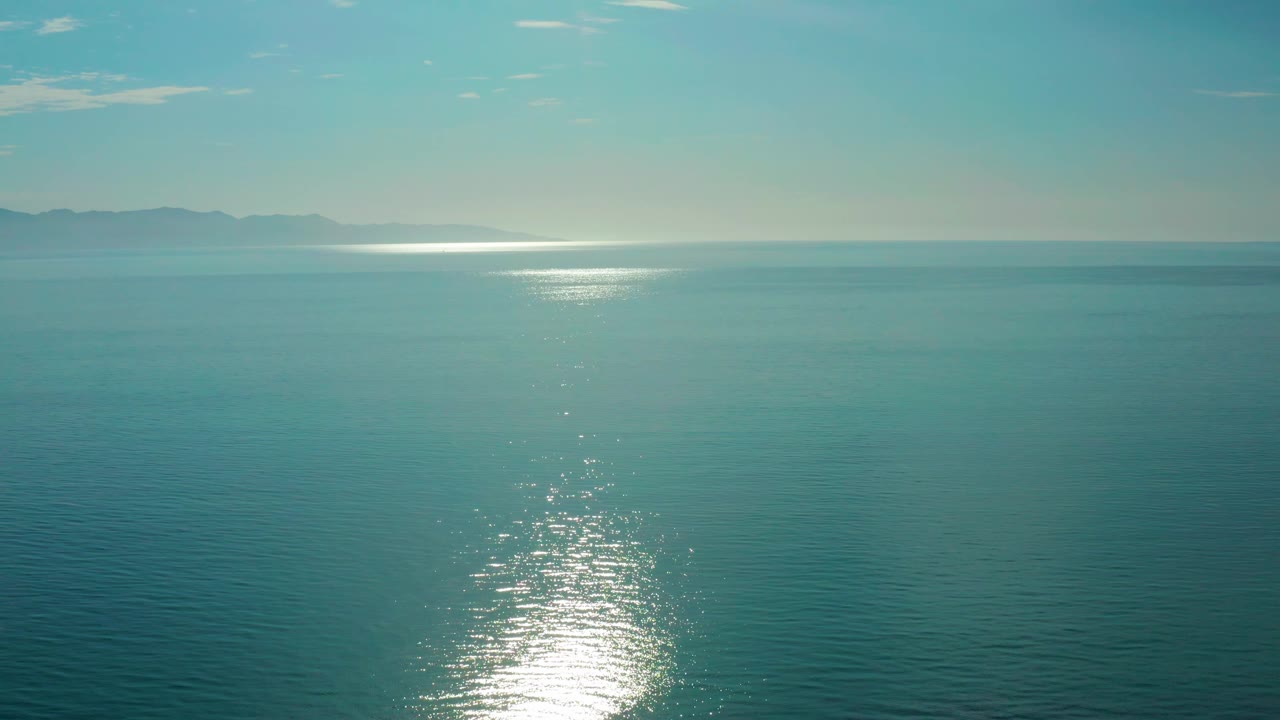 Mid-clip view at El Sargento La Ventana, Mexico, with calm blue-green ocean texture, a bright oval of reflected sunrise, distant hills along the upper left horizon, and no visible people.