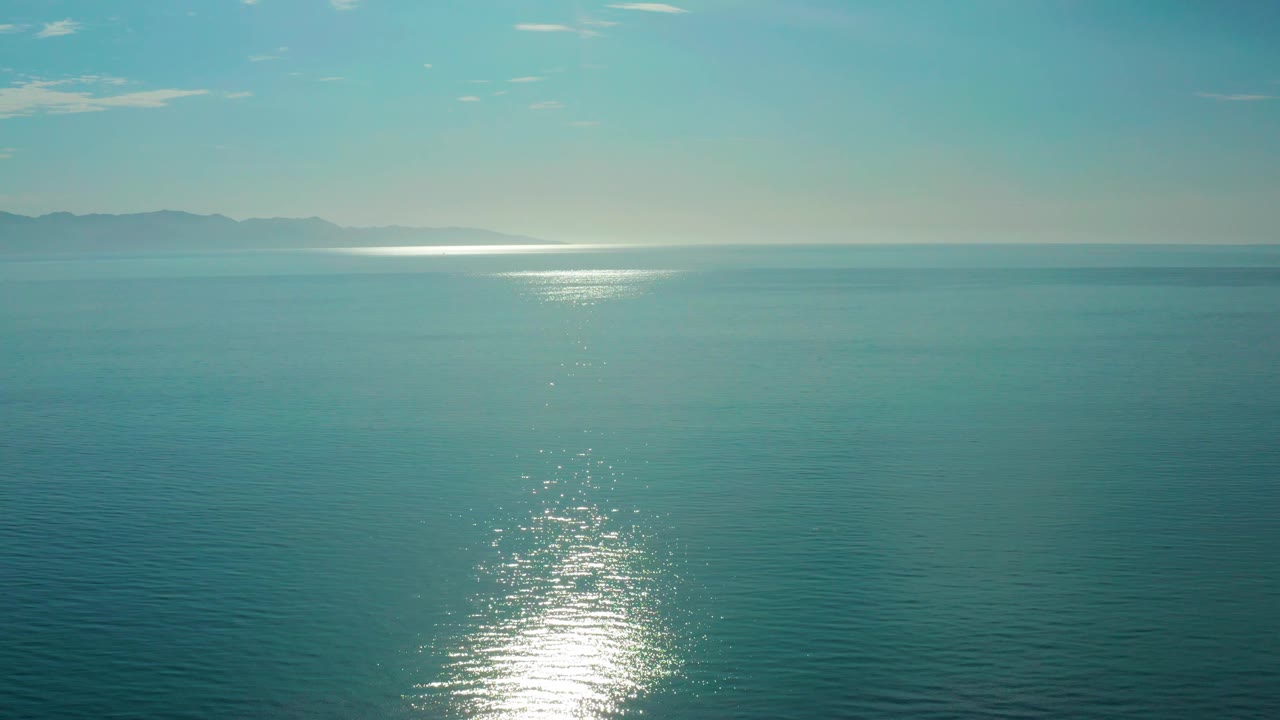 Frame at 0:03 over El Sargento La Ventana, Mexico, with the sea filling nearly the whole view, sunrise glare lower in frame, low mountains across the distant horizon, and no visible people.