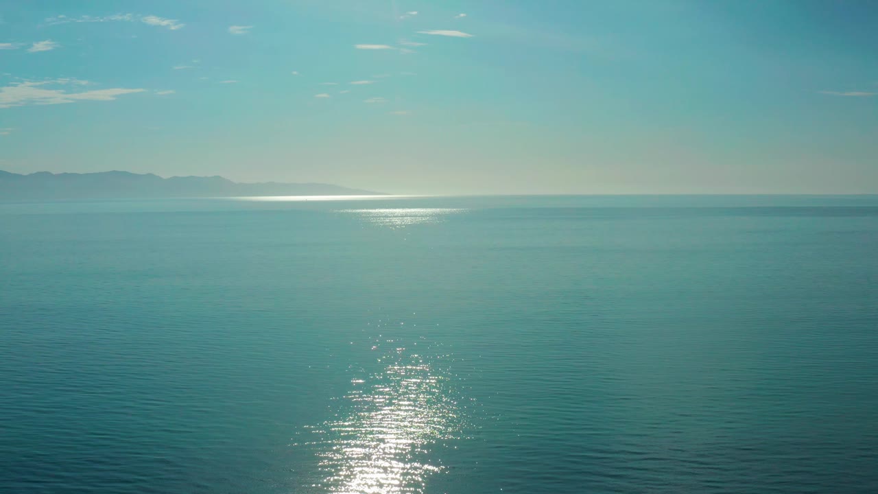 Blue sea surface in El Sargento La Ventana, Mexico, with a vertical patch of sun sparkle lower in frame and low landforms across the far horizon, no visible people.