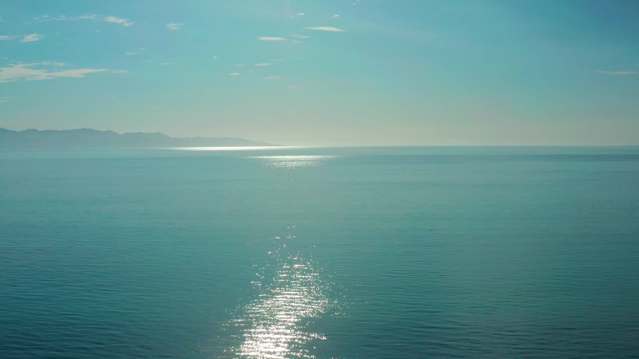 Calm ocean in El Sargento La Ventana, Mexico, with the sunlight patch low and left of center, hazy land at the horizon, and no visible people.