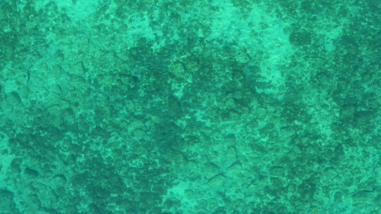 Straight-down aerial view of turquoise ocean surface and mottled bottom patterns, with no boats, shoreline, or visible people.