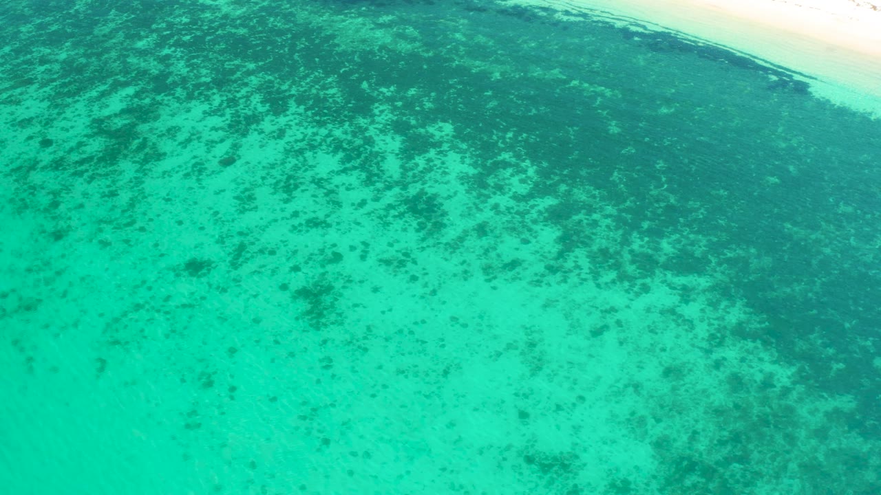 Aqua shallows fill the frame at El Sargento La Ventana, Mexico, with mottled dark reef or sea grass shapes below the surface and a thin beach strip near the top, no visible people.