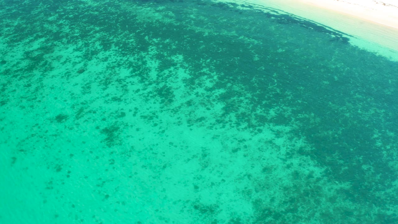 Aerial coastal frame at El Sargento La Ventana, Mexico, showing turquoise water crossed by darker submerged growth and a narrow bright sand margin near the upper right, no visible people.
