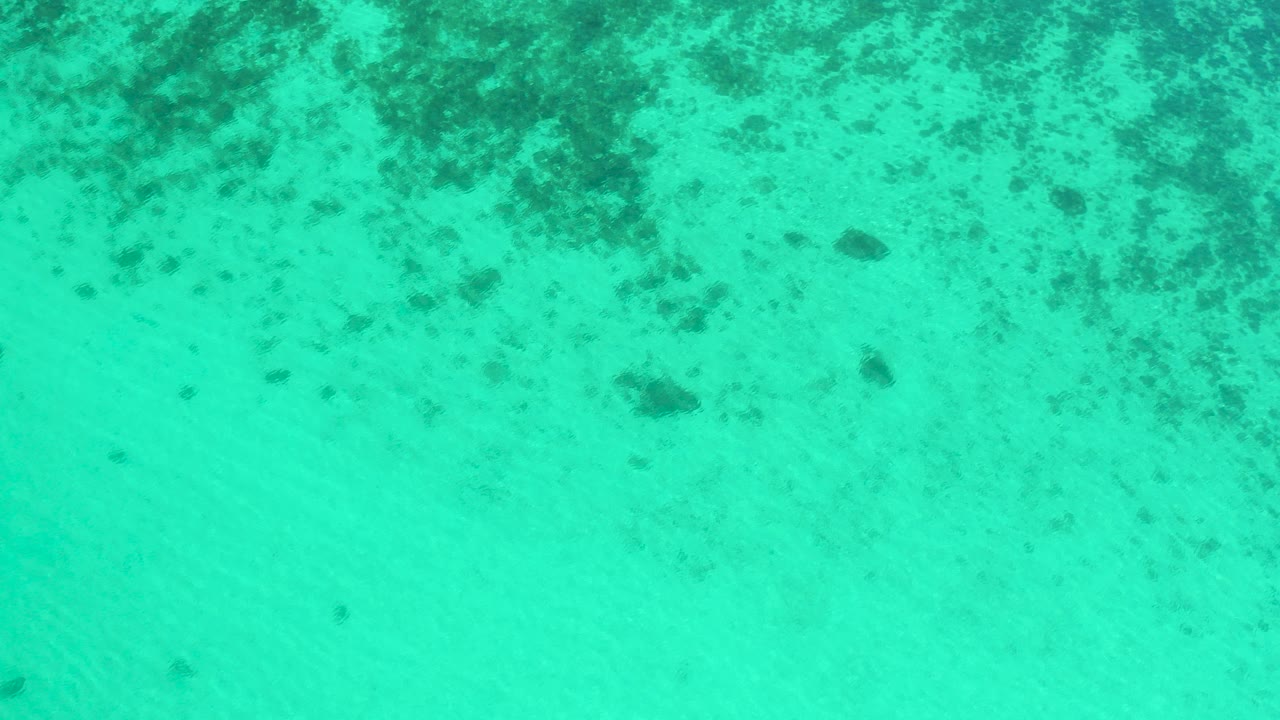Overhead view of turquoise shallow sea in El Sargento La Ventana, Mexico, with pale sand, scattered dark seabed patches, and no visible people.