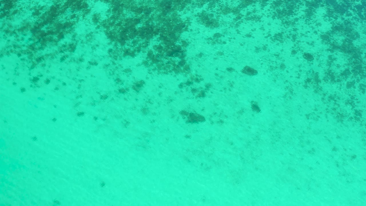Overhead aerial view of shallow turquoise sea in El Sargento La Ventana, Mexico, with a larger dark seabed patch on pale sand and no visible people.