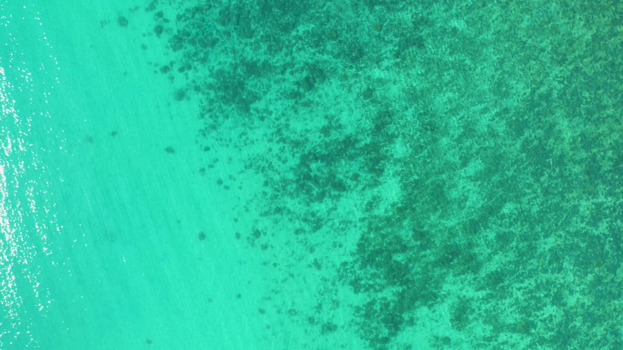 Top-down aerial view of turquoise ocean in El Sargento La Ventana, Mexico, with a broad dark underwater patch filling much of the upper half and no visible people.