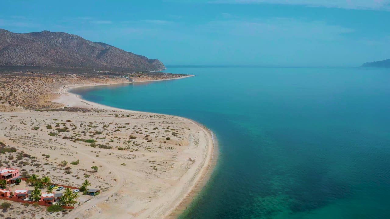 Aerial frame over El Sargento La Ventana, Mexico, showing the crescent beach more centered, turquoise shallows along the sand, dry desert ground, mountain slopes, and no visible people.
