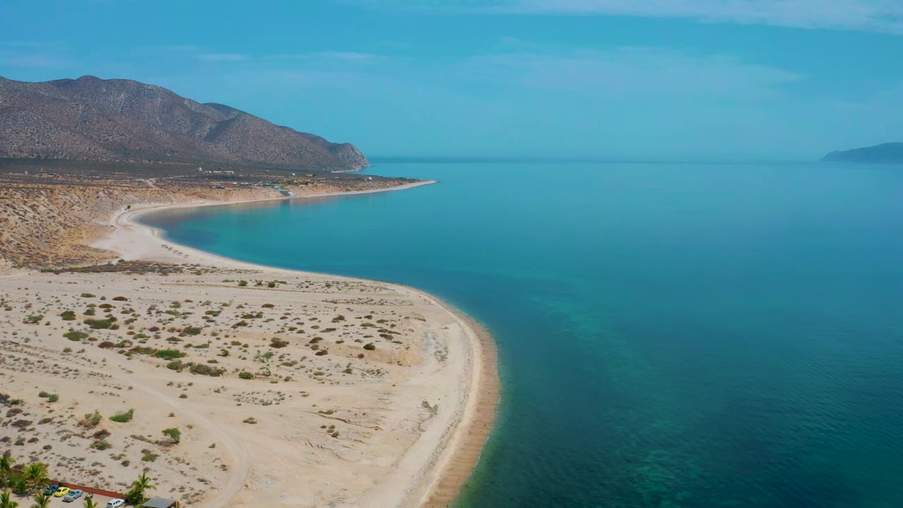 Aerial view over El Sargento La Ventana, Mexico, with desert scrub in the lower frame, a bright beach edge, blue-green water to the right, and no visible people.