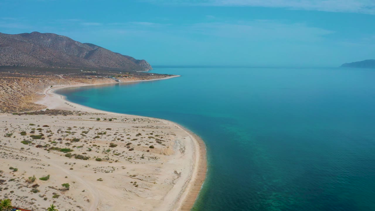 A high drone frame from El Sargento La Ventana, Mexico, showing the bay widening to the right, dry sandy land below, mountain slopes to the left, and no visible people.