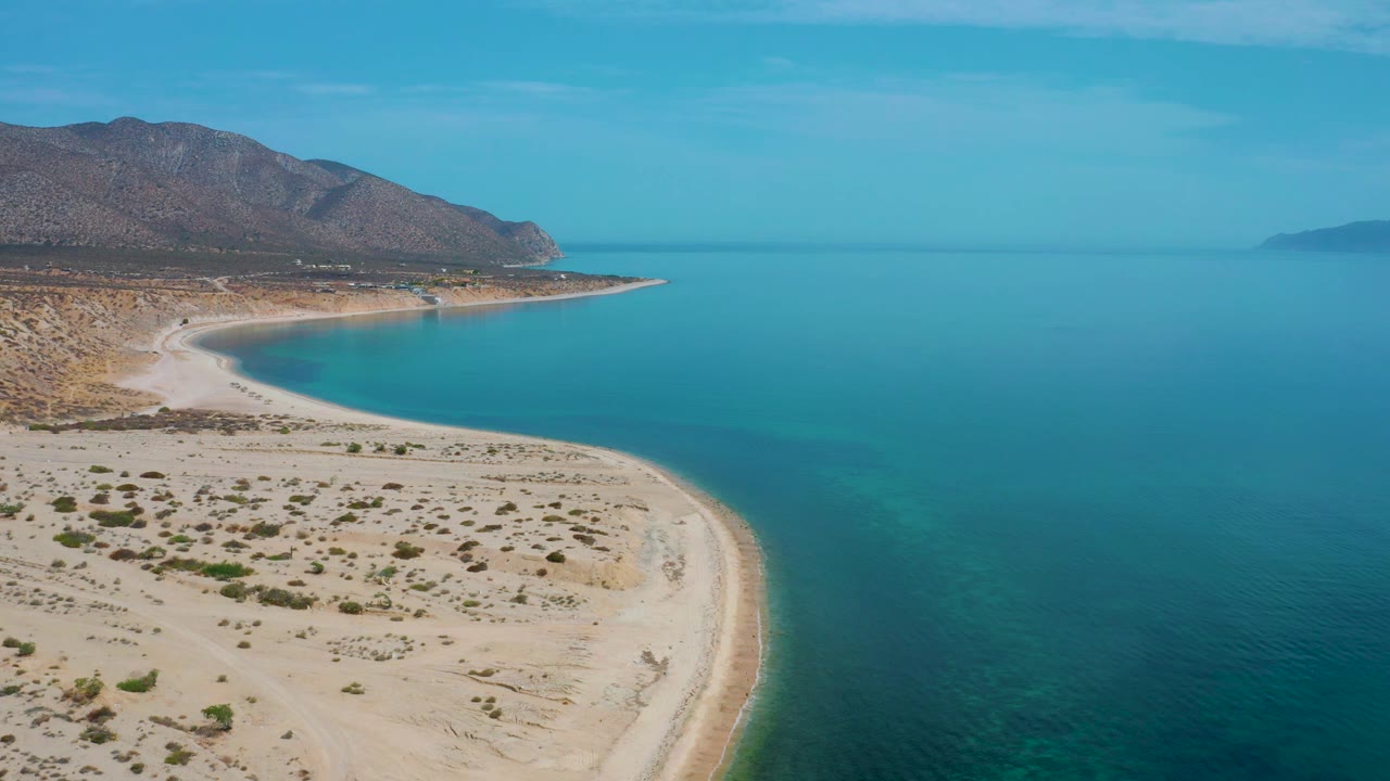 El Sargento La Ventana aerial frame with pale sand and scrub across the foreground, turquoise shallows along the shore, open ocean beyond, and no visible people.