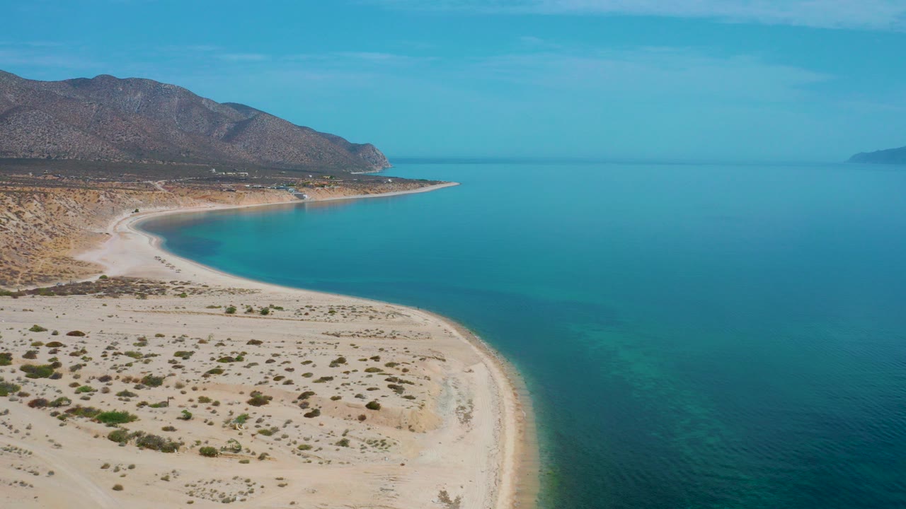 Aerial view of curved sandy shoreline, turquoise sea, desert scrub, and mountains.