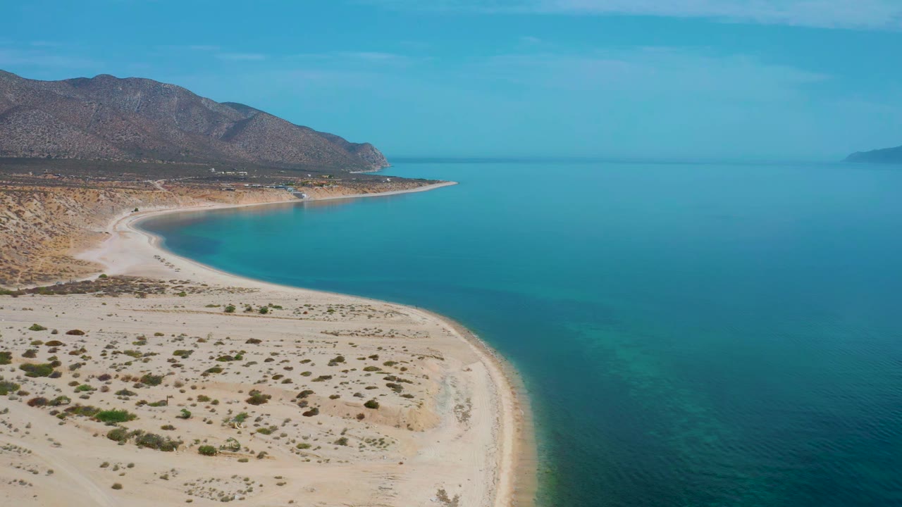 Aerial view in El Sargento La Ventana, Mexico, with the beach arc slightly farther through the frame, calm blue water to the right, rugged hills to the left, and no visible people.