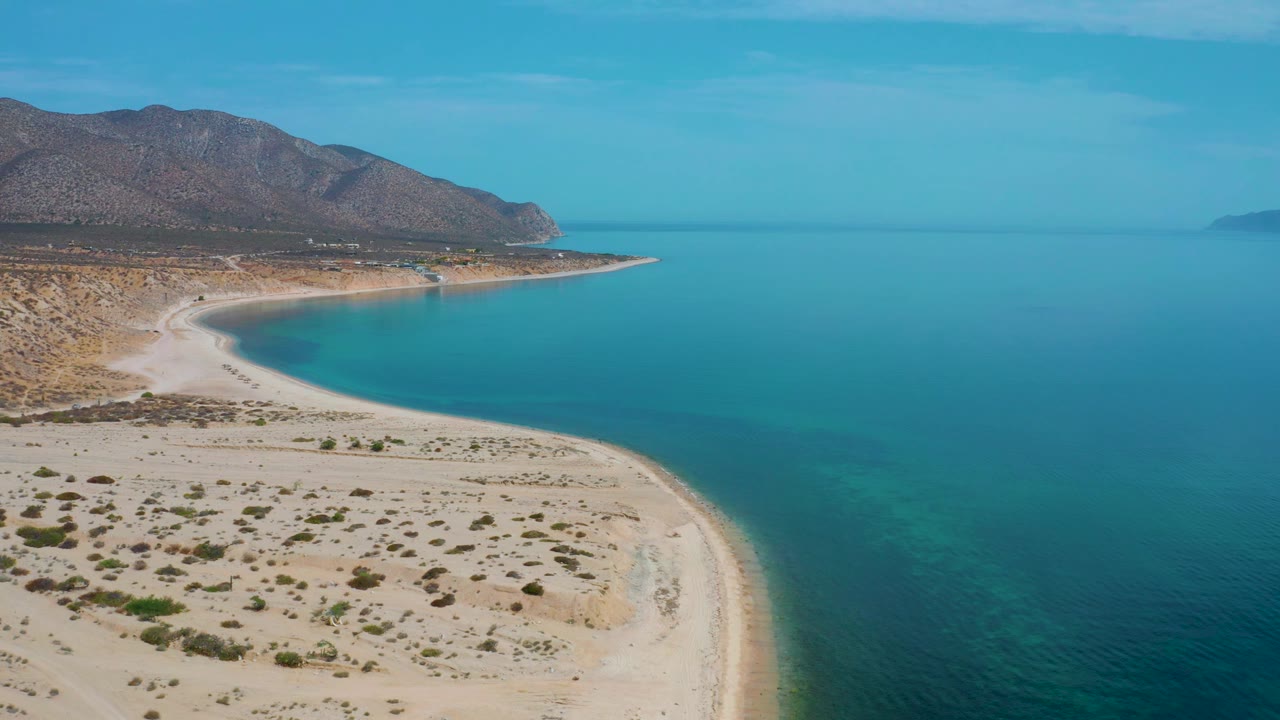 High view over El Sargento La Ventana, Mexico, where pale sand and desert brush fill the lower frame, clear water bands run along the shore, mountains sit beyond the bay, and no visible people appear.