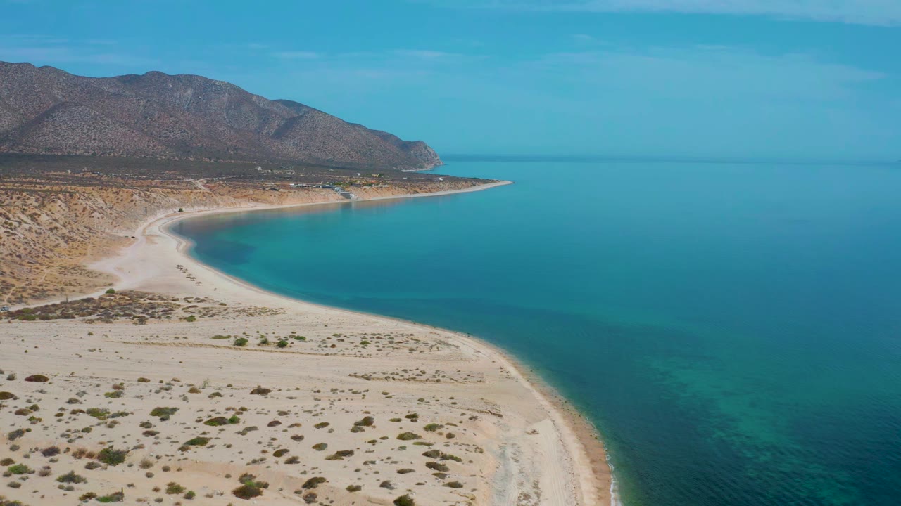 High coastal view with pale sand, blue-green water, scrubland, and mountains.