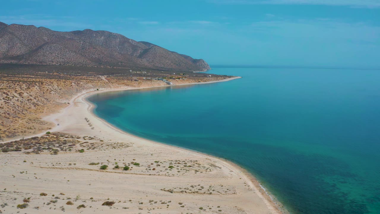 Wide aerial frame of El Sargento La Ventana, Mexico, showing the sandy cove slightly closer, calm blue-green water, scrubland, mountains, and no visible people.